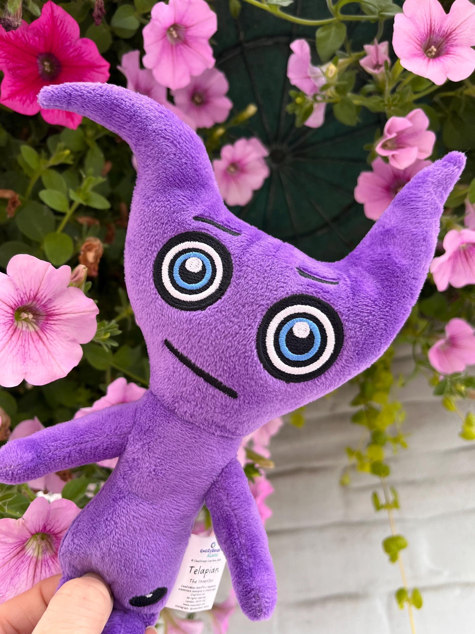 CwaZyBean Aliens Purple plush toy Telapian with large eyes from CwaZyLeigh CreaTions held in front of pink flowers 